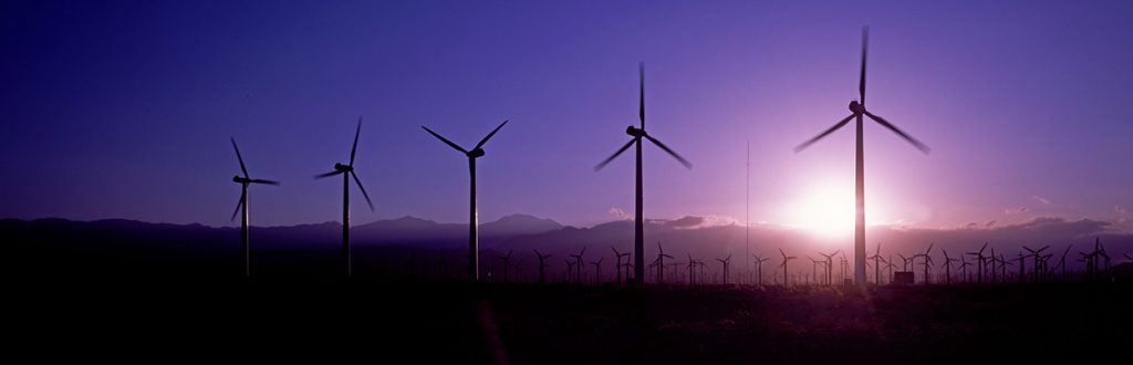 Mehrere Windkraftanlagen stehen auf einem Feld. Im Hintergrund geht die Sonne unter und verfärbt den Himmel in lila.