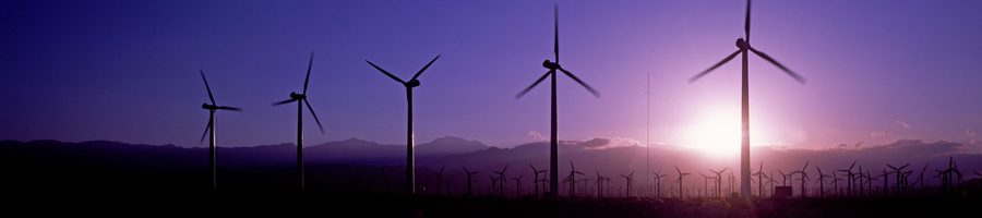 turbines-Slider Mehrere Windkraftanlagen stehen auf einem Feld. Im Hintergrund geht die Sonne unter und verfärbt den Himmel in lila.