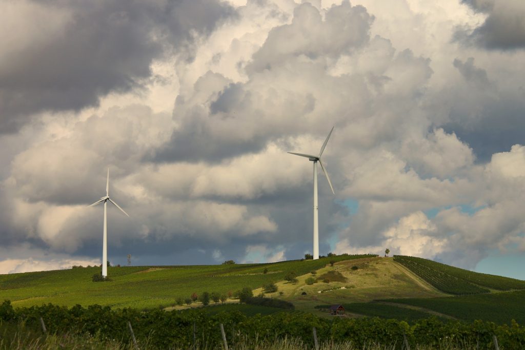 Zwei Windkraftanlagen stehen auf grün bewachsenen Berghügel. Der Himmel ist mit dicken Wolken bedeckt.