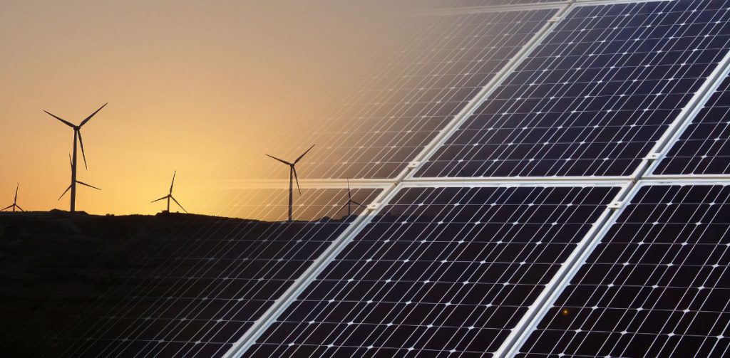 In der einen Hälfte stehen Windkraftanlagen auf einem dunklen Feld, im Hintergrund geht die Sonne unter. In der zweiten Hälfte geht die Kulisse der Windkraftanlagen über zu einer Fläche von Solaranlagen.