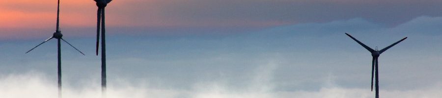 Windkraftanlage Drei Windkraftanlagen ragen aus Wolken in den Himmel, der durch den Sonnenuntergang einen orangen Streifen bekommt.