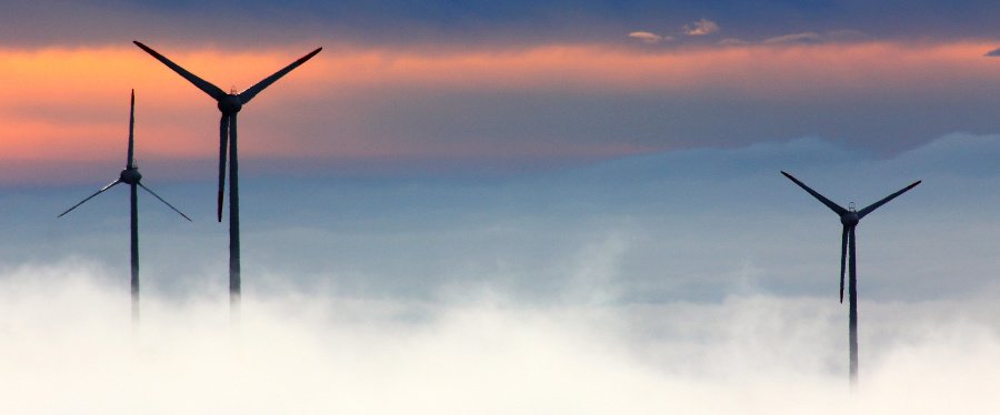 Drei Windkraftanlagen ragen aus Wolken in den Himmel, der durch den Sonnenuntergang einen orangen Streifen bekommt.
