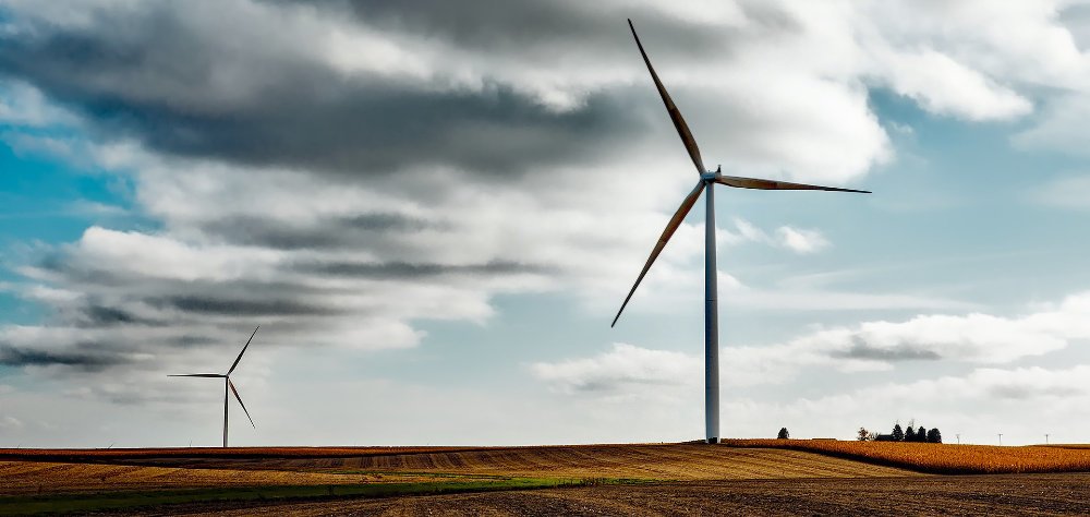 Eine Windkraftanlage im Vordergrund und eine weitere im Hintergrund stehen auf einem abgeernteten Getreidefeld. Der Himmel ist mit grauen Wolken bedeckt.
