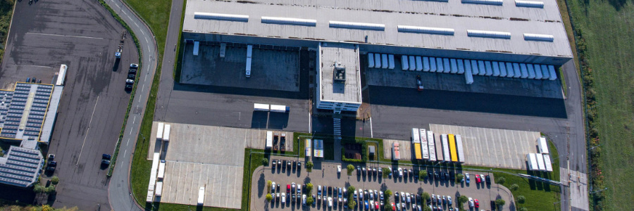 RealEstate_Logistik_VelpeDJI_0018_Web-Beitrag Aus der Luft ist eine große Logistikhalle zu sehen. Davor ein großer Parkplatz mit Autos.